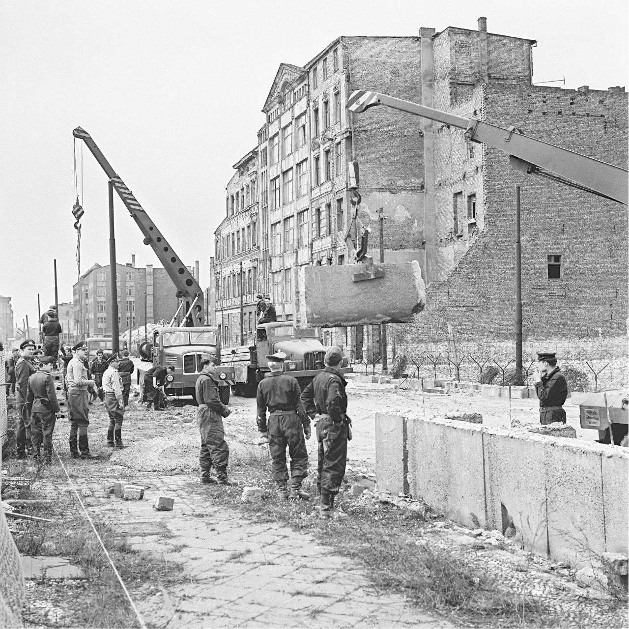 Soldaten der Volkspolizei der DDR beim Errichten der Mauer in der Zimmerstrasse in Berlin. Im  Vordergrund steht schon ein Stück Mauer. Im Hintergrund zwei Kräne und Häuserblöcke; einer der Kräne hebt gerade ein Stück Mauer.