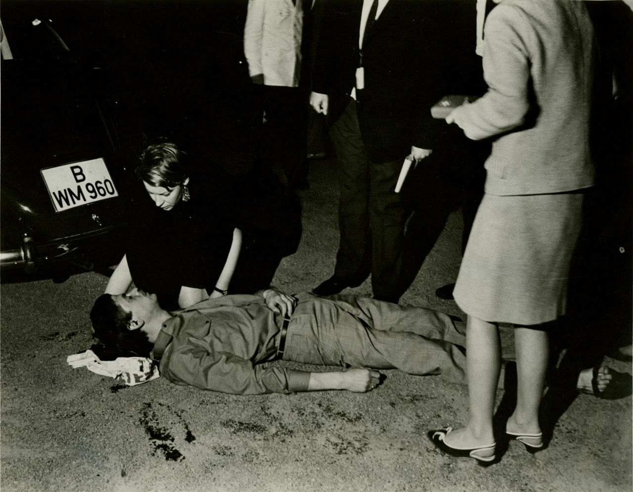 Benno Ohnesorg liegt schwer verletzt auf dem Rücken am Boden; unter seinem Kopf ein Tuch. Eine Frau an seinem Kopf kniend beugt sich über ihn. Zwei weitere Personen stehen um ihn herum. Im Hintergrund das Heck eines Autos mit dem Kennzeichen B WM 960. 