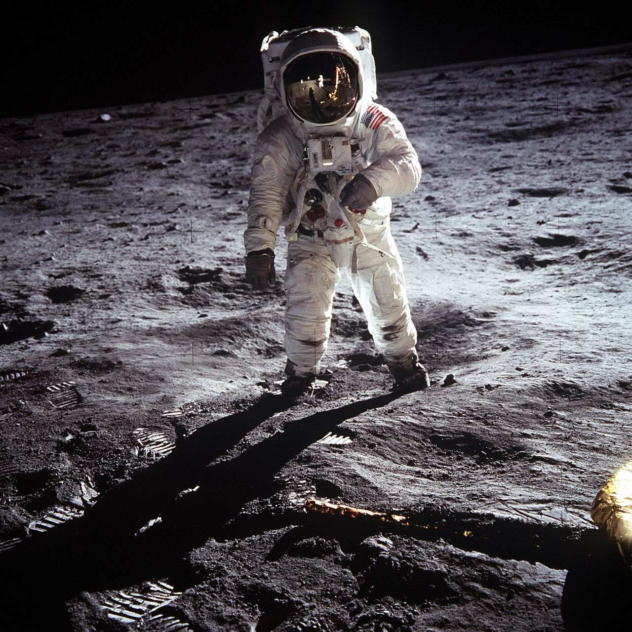 Buzz Aldrin geht auf der unebenen Mondoberfläche. Er trägt einen weißen Raumanzug mit USA-Flagge auf der linken Schulter und mit einem nahezu kugelförmigen Helm.