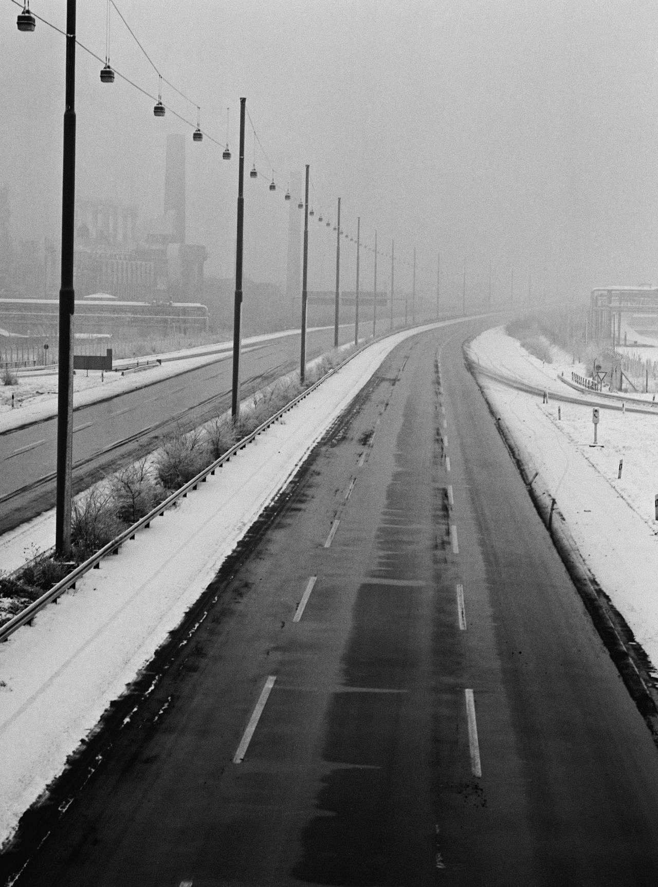 Schwarzweiß-Foto einer autofreien Autobahn. Je Fahrtrichtung drei Spuren. Auffahrt von rechts. Mittel- und Randstreifen sind schneebedeckt. Auf dem Mittelstreifen Straßenlaternen. Hintergrund nebelverhangen. Links neben der Autobahn Industrieanlagen.