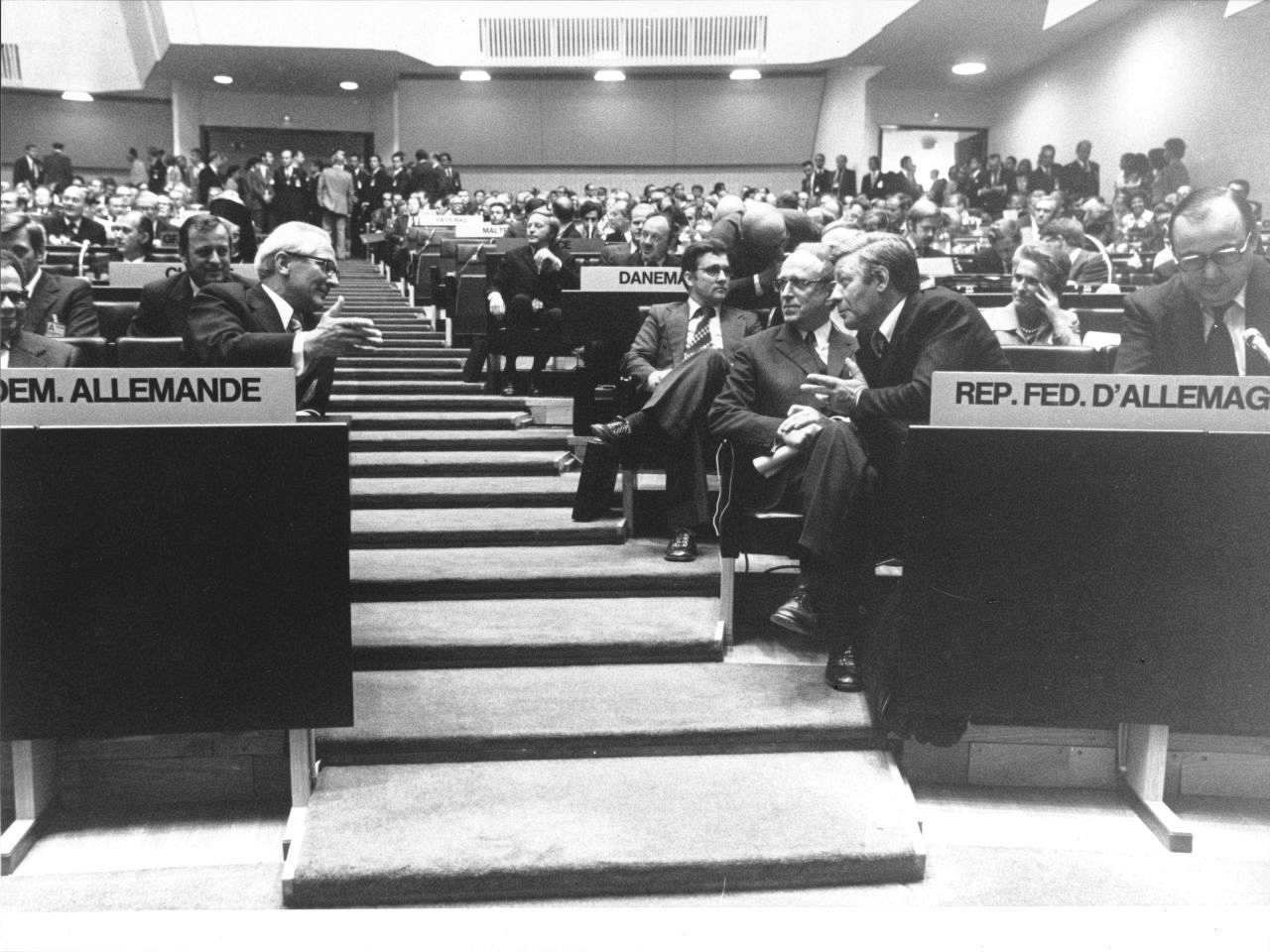 Schwarzweiß-Foto des KSZE-Konferenzsaals. Mittig Treppe hinauf zum Ausgang. Beidseitig davon Rang-Bestuhlung. In der ersten Reihe durch die Treppe voneinander getrennt, sitzen rechts Erich Honecker und links Helmut Schmidt, miteinander im Gespräch.