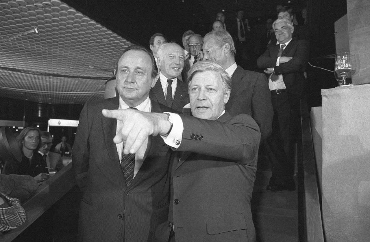 Schwarzweiß-Foto im Inneren eines Gebäudes. Mehrere Personen stehen auf einer Treppe. Vorne links Genscher und rechts Helmut Schmidt, beide mit gleicher Blickrichtung.  Schmidt zeigt mit dem Finger in Blickrichtung. Dahinter Scheel und Brandt im Gespräch.
