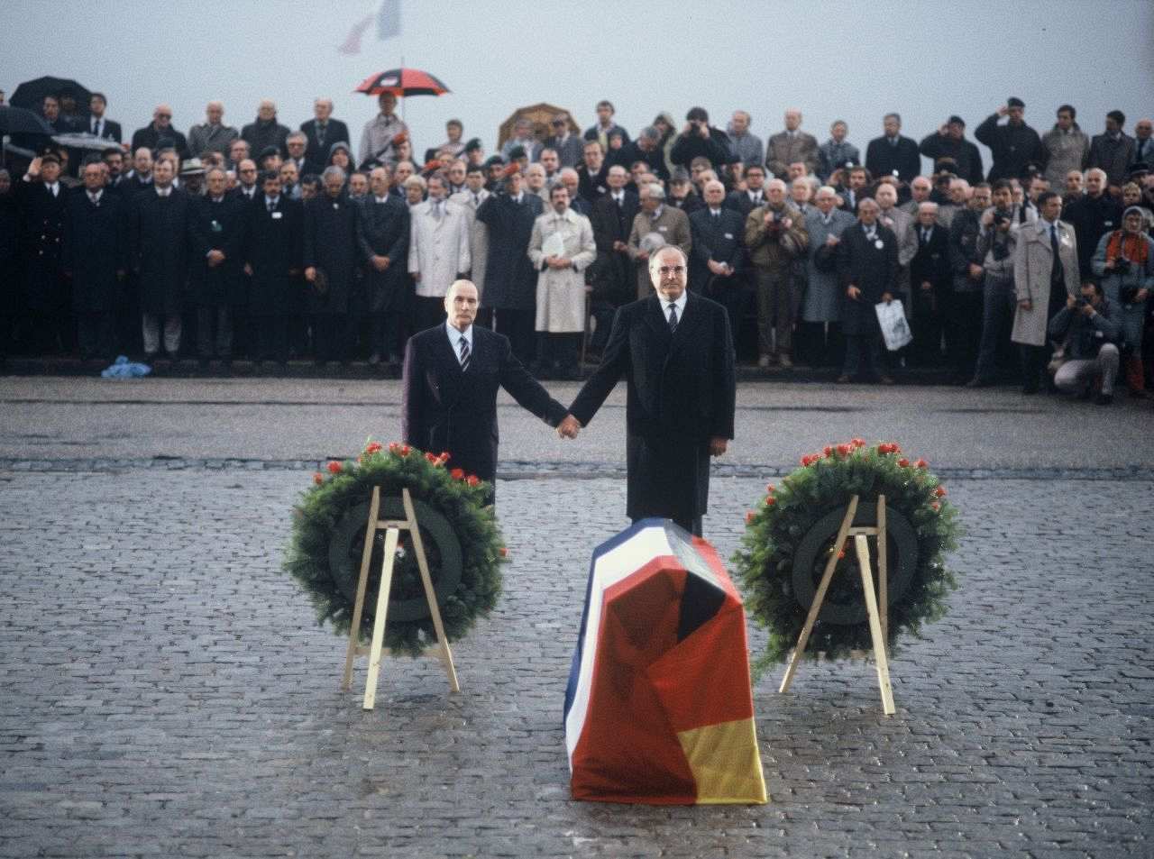 Farbfoto, Francois Mitterrand und Helmut Kohl stehen Hand in Hand auf Kopfsteinpflaster vor zwei aufgestellten Kränzen und einem mit deutscher und französischer Fahne abgedeckten sargartigen Gebilde. Im Hintergrund eine Menschenmenge, teils mit Schirmen. 