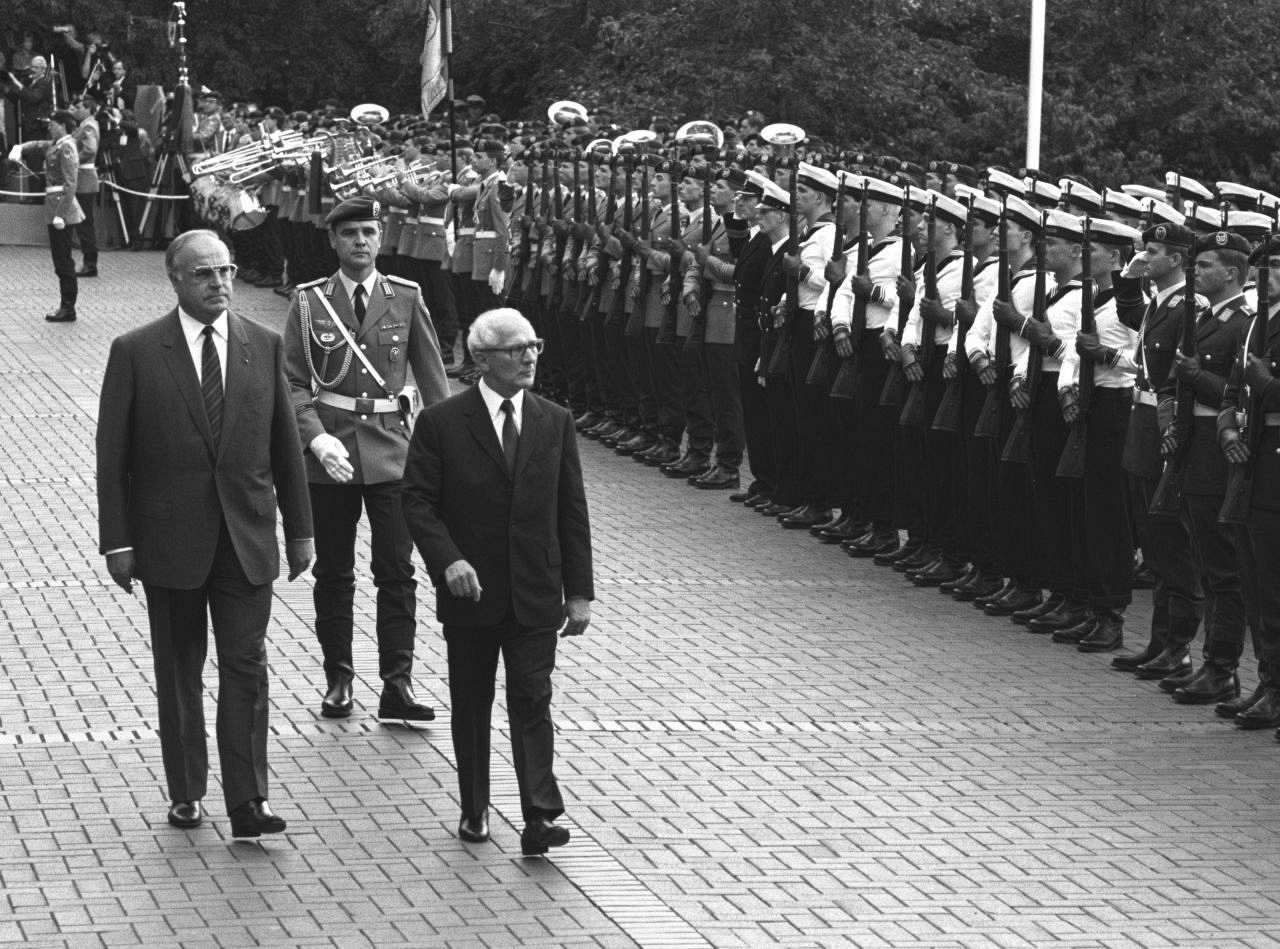 Schwarzweiß-Foto, Helmut Kohl (links) und Erich Honecker (rechts), gefolgt von einem Soldat, schreiten entlang einer militärischen Ehrenformation, bestehend aus Musikkorps und Kompanien verschiedener Teilstreitkräfte. Die Soldaten präsentieren das Gewehr.