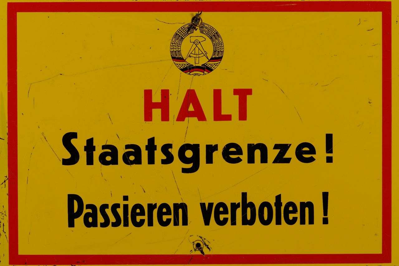 Rechteckiges, gelbes Kunststoffschild, rot gerahmt, oben Staatswappen der DDR (Hammer und Zirkel im Ährenkranz), darunter die rote Aufschrift HALT. Darunter in schwarzer Schrift: Staatsgrenze! Passieren verboten!