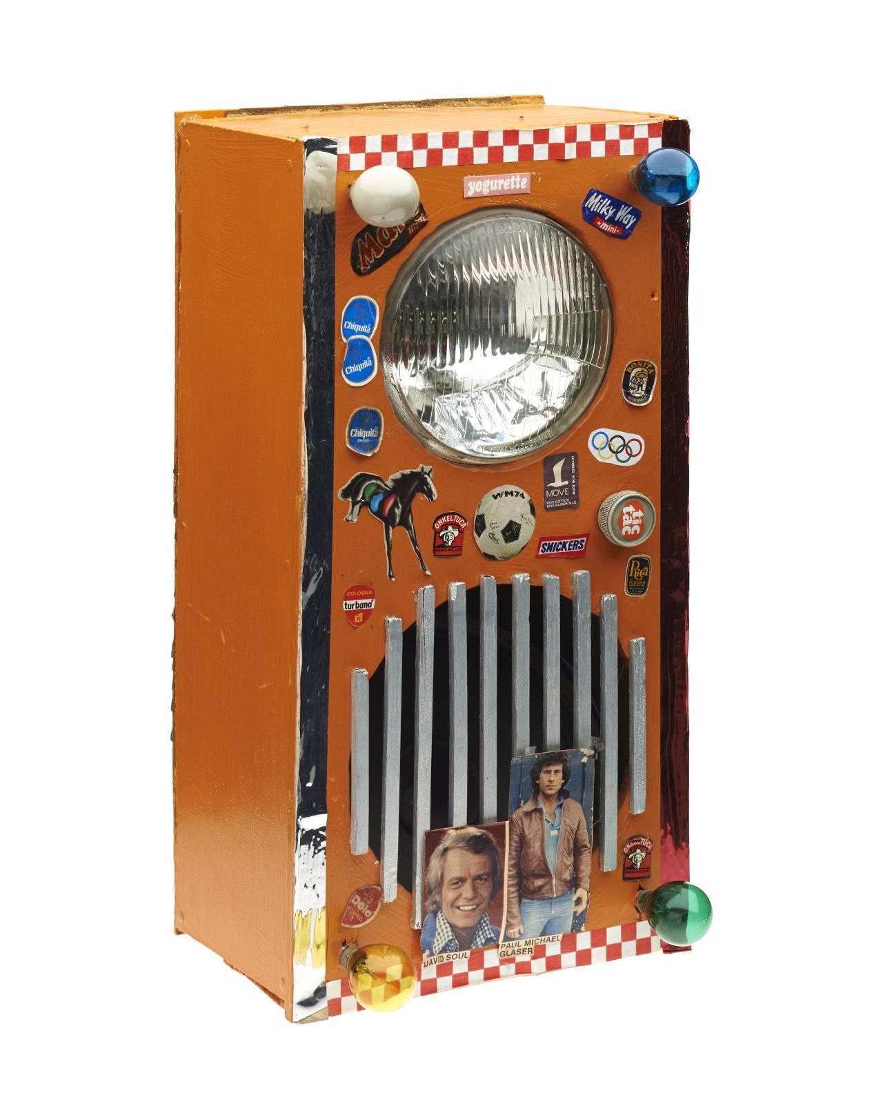 Hochrechteckiger Sperrholzkasten, orange lackiert. Eine Aussparung für den Lautsprecher mit einem silberfarbenem Gitter. Darüber eine weiße Autoleuchte. Verzierung: farbige Glühlämpchen, diverse westliche Produktaufkleber, Bilder von Starsky und Hutch.