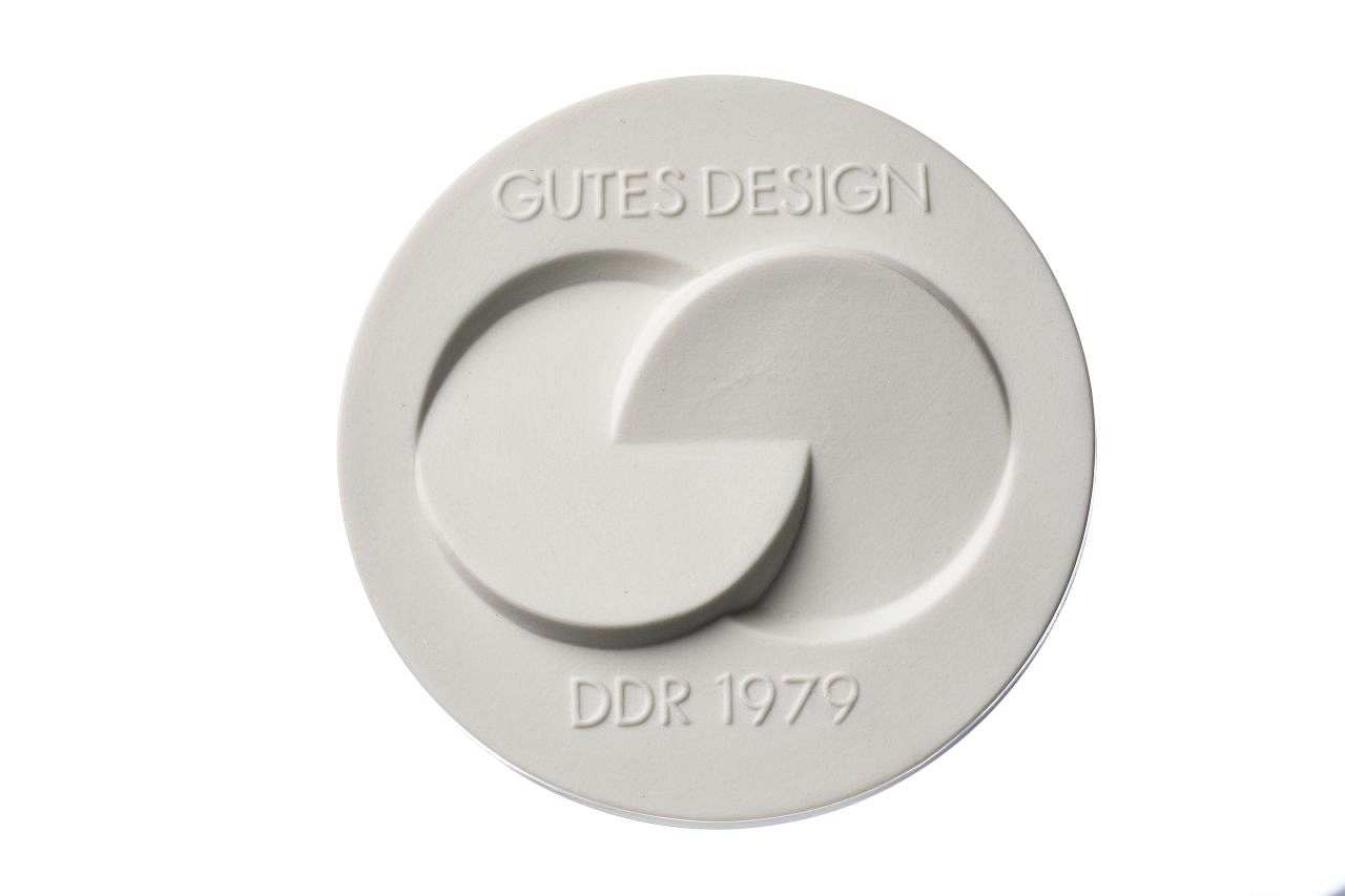 Weiße Porzellanmedaille, Vorderseite mittig erhabenes Signet, oben Gutes Design, unten DDR 1979, Rückseite DDR-Staatssymbol sowie Schwertermarke der Meißner Porzellanmanufaktur, weißes quadratisches Etui, innen mit weißem und blauem Stoff bezogen.