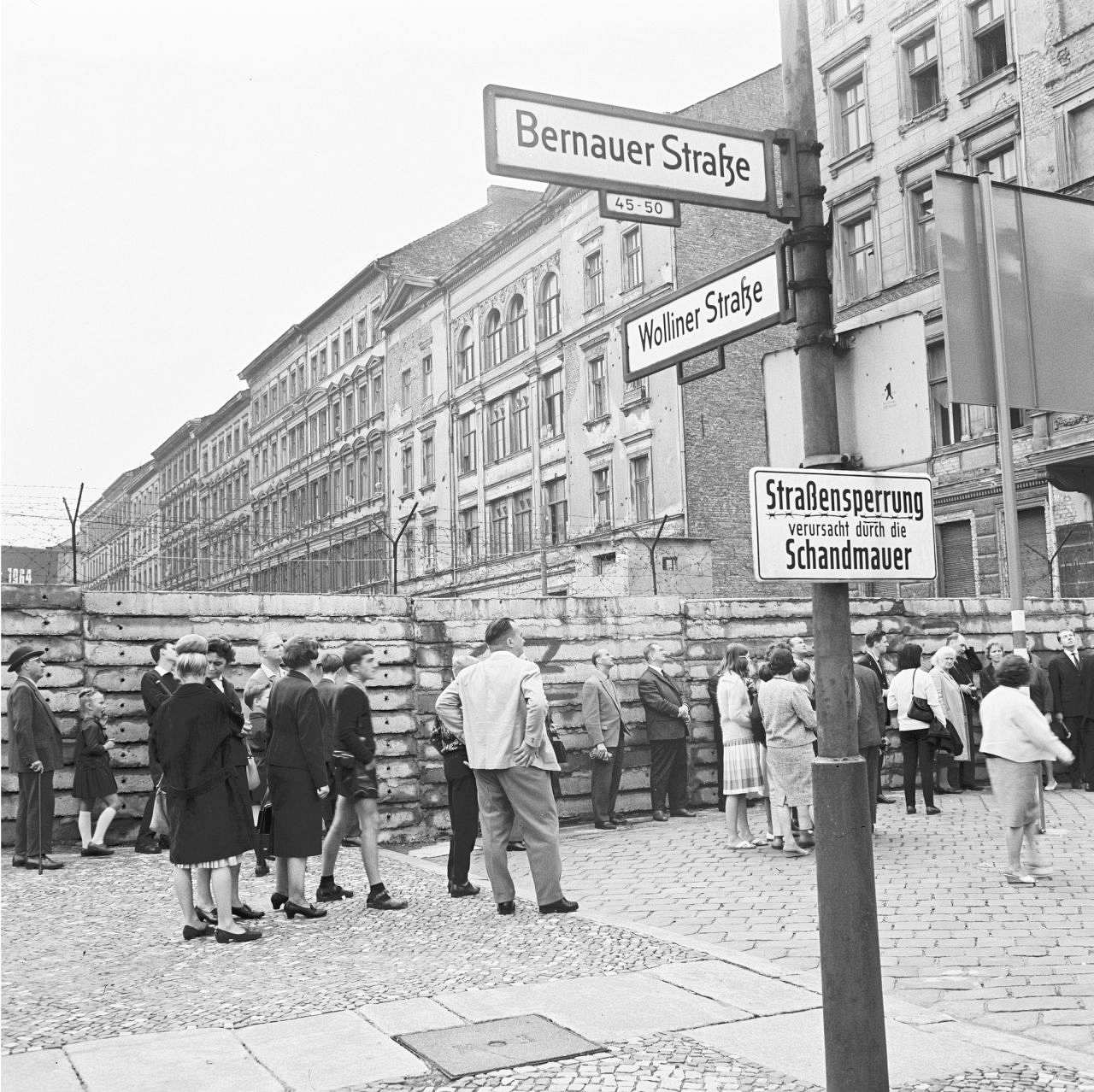 Schwarz-weiße Aufnahme: Personen stehen entlang der Berliner Mauer auf der Bernauer Straße, Höhe Wolliner Straße in West-Berlin. Rechts im Vordergrund Schild mit der Aufschrift: Straßensperrung verursacht durch die Schandmauer.