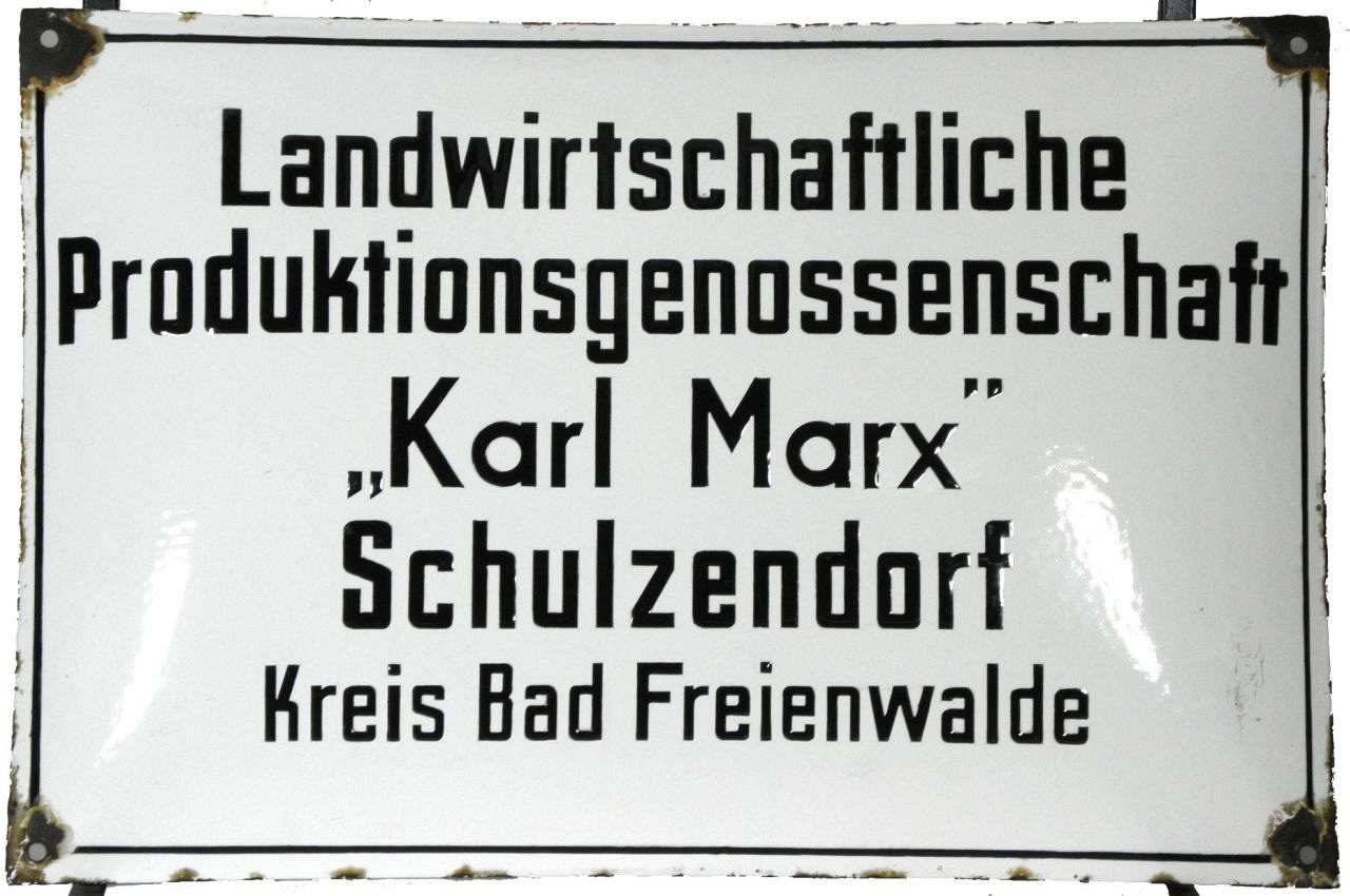 Gewölbtes weißes Schild mit schwarzer Beschriftung: Landwirtschaftliche - Produktionsgenossenschaft - Karl Marx - Schulzendorf - Kreis Bad Freienwalde, schwarz umrandet. In den Ecken Befestigungslöcher.