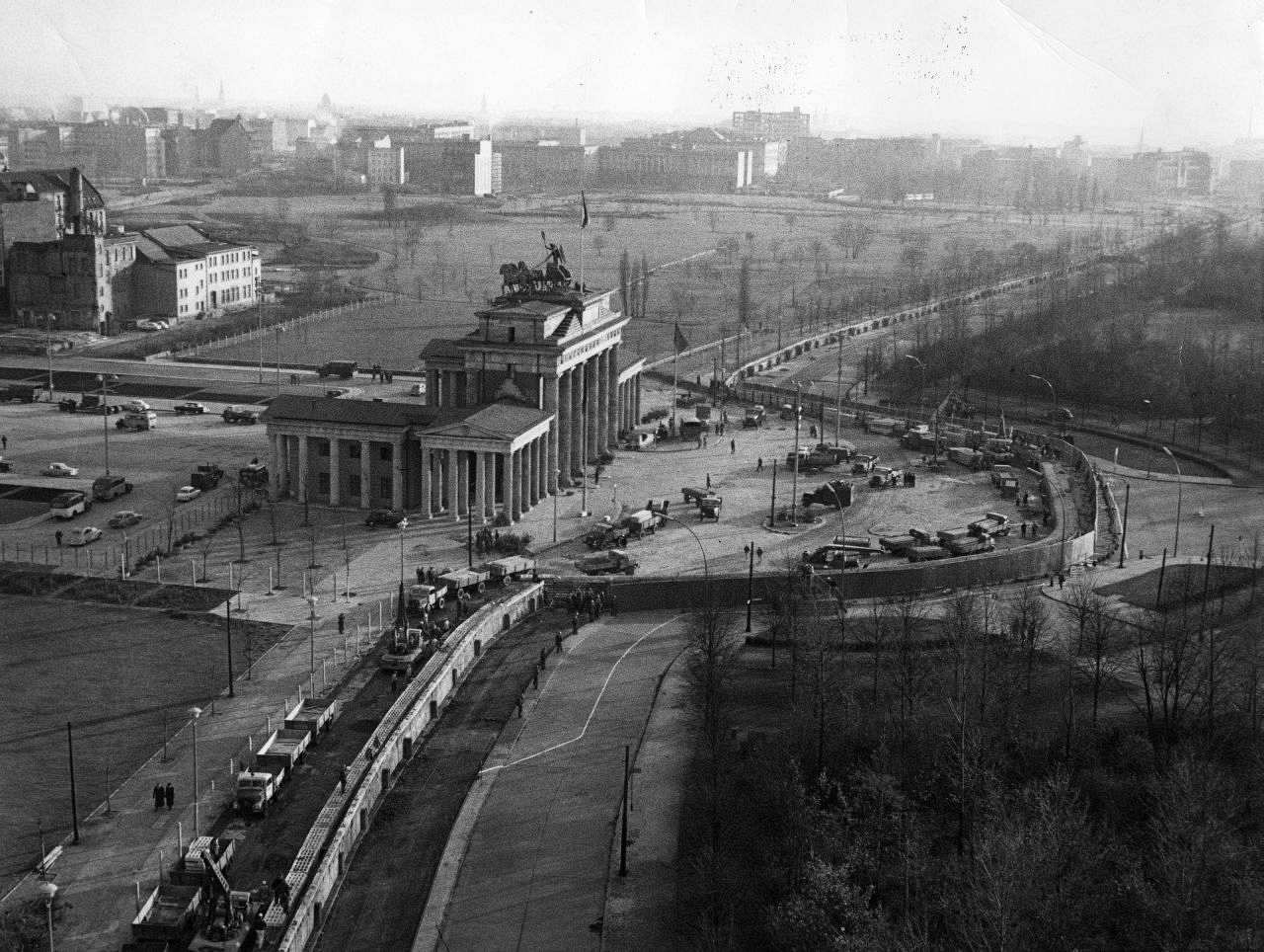Seitlicher Blick von oben auf das Brandenburger Tor im Zentrum des Bildes. Eine Mauer läuft vertikal durch das Bild mit einer großen Ausbuchtung um das Tor. Links an der Mauer viele Fahrzeuge, im Hintergrund Freifläche und Gebäude; rechts davon Bäume.