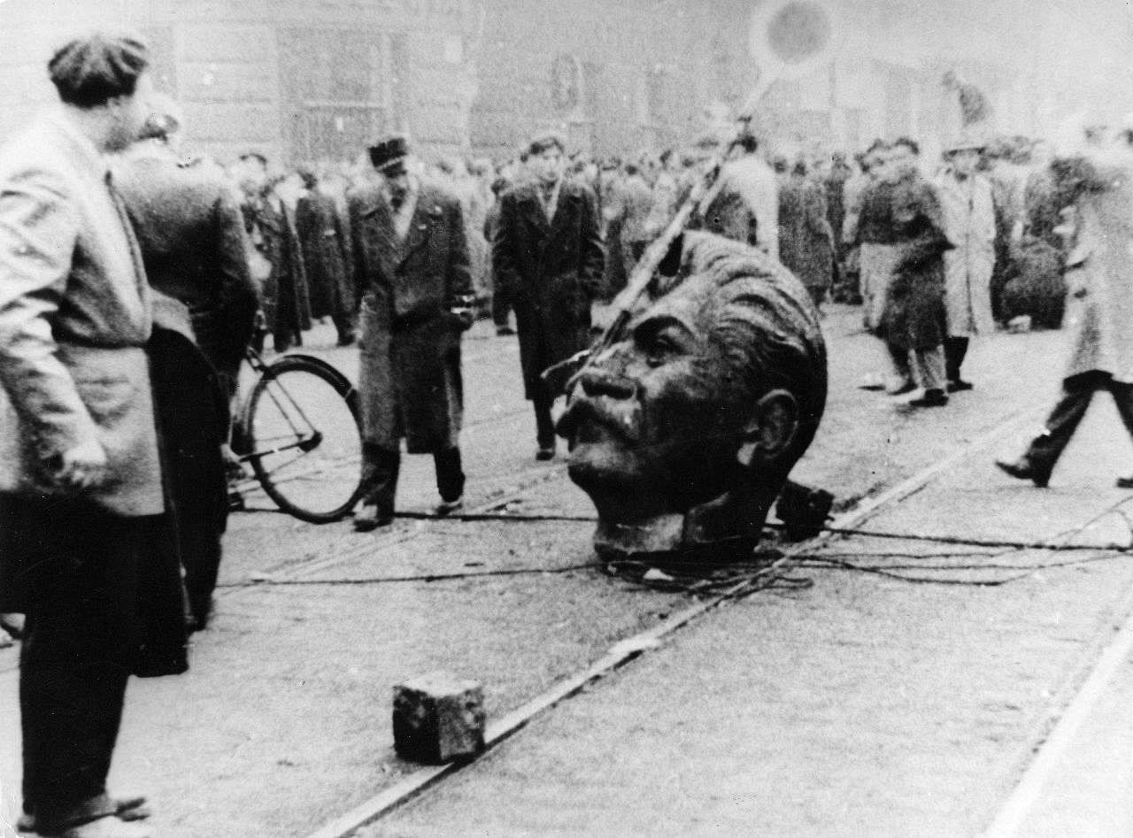 Bildmitte: abgetrennter Kopf eines Stalin-Denkmals liegt auf Straße sowie Pflasterstein und Kabel, im Hintergrund: Menschenmenge.