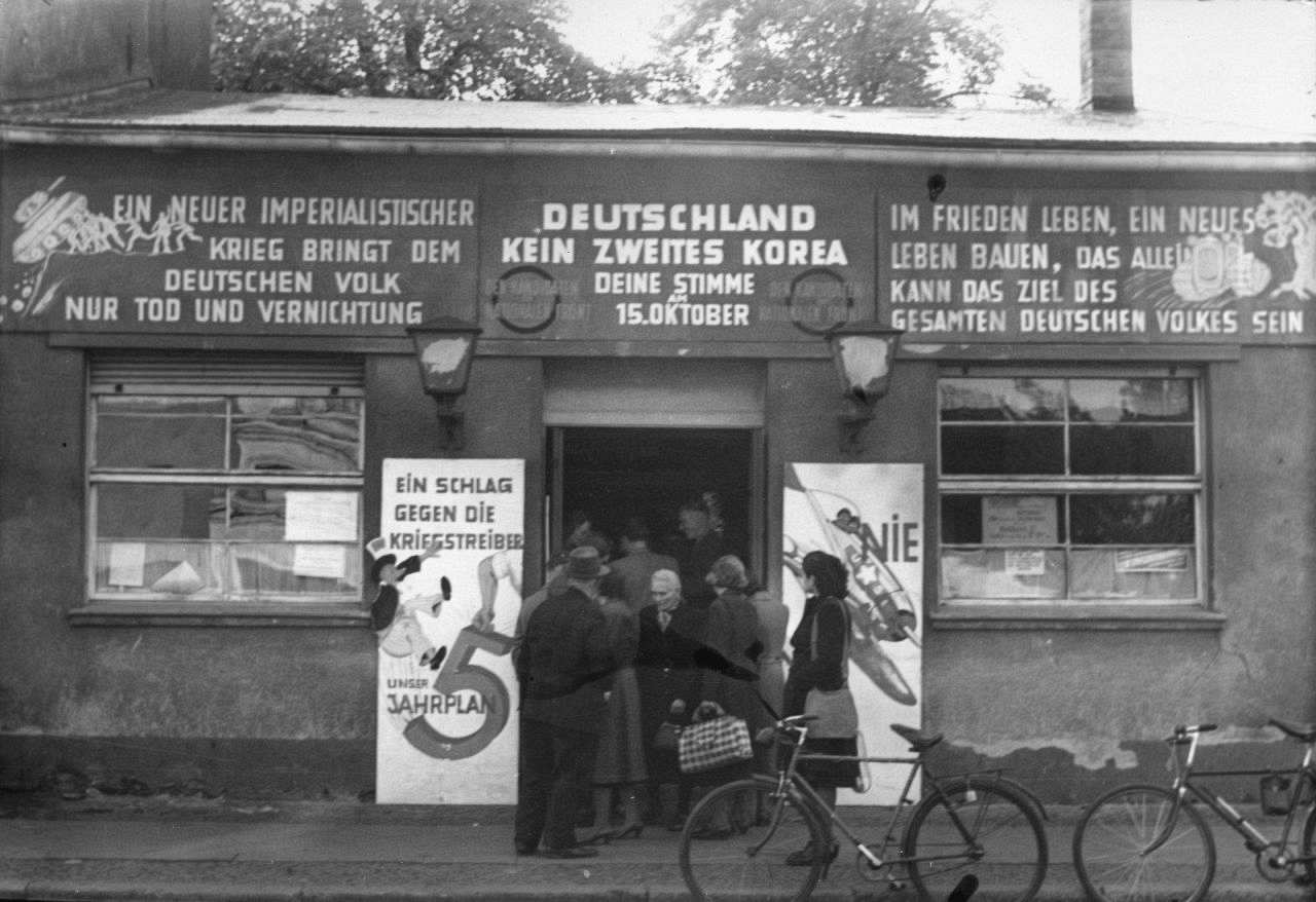 Schwarzweißfoto; flaches Gebäude, vor und im Eingang wartende Menschen. An der Fassade hängen Schilder mit Wahlkampfparolen. Zwei Herrenfahrräder sind vor dem Bürgersteig abgestellt.