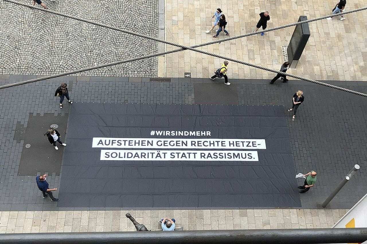 Das Banner mit der Aufschrift „#wirsindmehr. Aufstehen gegen rechte Hetze - Solidarität statt Rassismus.“ hängt beim „Wir sind mehr“-Konzert am 3. September 2018 in Chemnitz im Hintergrund auf der Bühne. Nach einem tödlichen Messerangriff am Rande des Chemnitzer Stadtfestes kommt es zu rechtsextremen Ausschreitungen, bei denen tatsächliche oder vermeintliche Migranten verfolgt und angegriffen werden. Als Reaktion darauf organisiert die Chemnitzer Band Kraftklub mit Unterstützung des Chemnitzer Stadtmarketings ein kostenloses Konzert als Gegendemonstration.