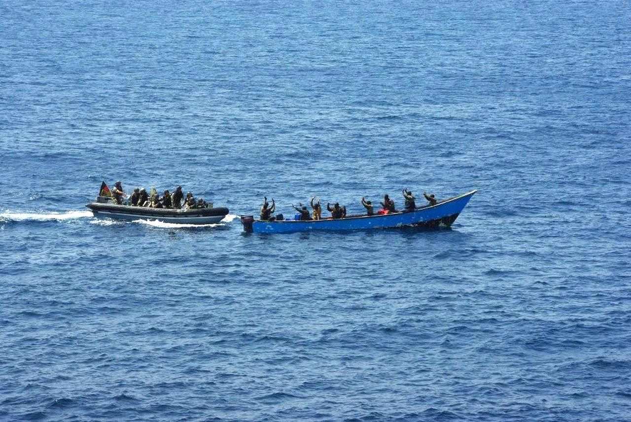 Das Foto zeigt zwei Boote mitten im Meer. Das Wasser ist tiefblau und sehr ruhig. Ein Boot voller Soldaten fährt an ein Piraten-Boot heran. Acht Piraten sind zu erkennen, die alle die Hände hochheben. Am Bundeswehr-Boot ist eine deutsche Fahne zu erkennen. Die Soldaten tragen alle Uniformen und Ausrüstung.