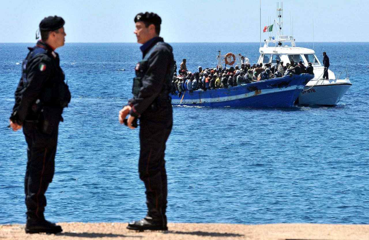 Zwei Soldaten am Meeresufer in Lamepdusa stehen sich gegenüber und unterhalten sich. Im Hinterrund sieht man auf dem blauen, stillen Meer ein Boot ankommen. Das Boot ist voll von Flüchtlingen, die sehr eng nebeneinander sitzen und es ist sichtbar zu schwer beladen.