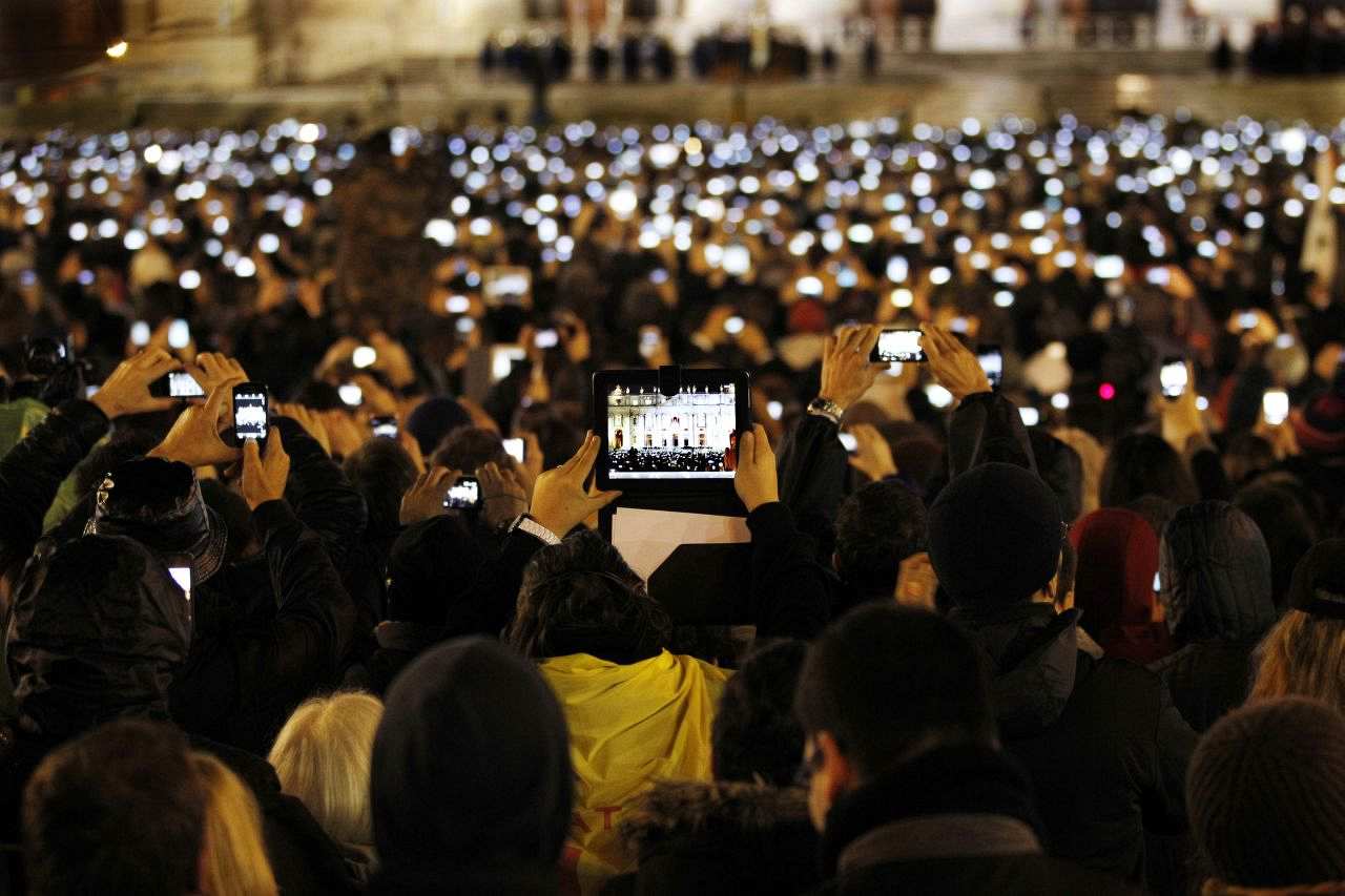 Eine dichte Menschenmenge am Abend steht auf dem Petersplatz vor dem Petersdom im Vatikan. Im Hintergrund gerade noch zu erkennen: Der Balkon des Petersdom auf dem der neu gewählte Papast Franziskus steht. Da alle Besucher mit ihren Smartphones und Tablets Papst Franziskus fotografieren, leuchtet ein Lichtermeer auf.