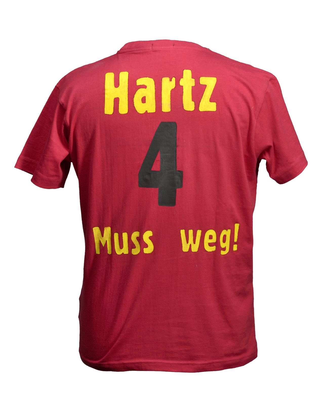 Rotes, klassisch geschnittenes Herren-T-Shirt mit rundem Halsausschnitt. Rückseite mit der Beschriftung analog zu Fußballtrikots: Oben steht Hartz, darunter die Zahl 4, darunter: Muss weg! Die Buchstaben sind gelb, die Zahl ist schwarz.