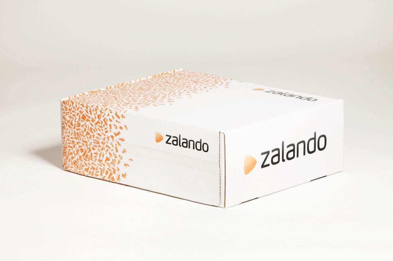 Zalando-Paket: Ein rechteckiger, weißer Karton. Auf allen drei sichtbaren Seiten ist das Zalando-Logo abgelichtet, ein hellorangenes Dreieck mit abgerundeten Ecken und schwarzer Schriftzug: zalando. Außerdem ist eine Hälfte des Kartons vollgesprenkelt mit kleinen, hellorangenen Grafiken von Schuhen und Kleidungsstücken.