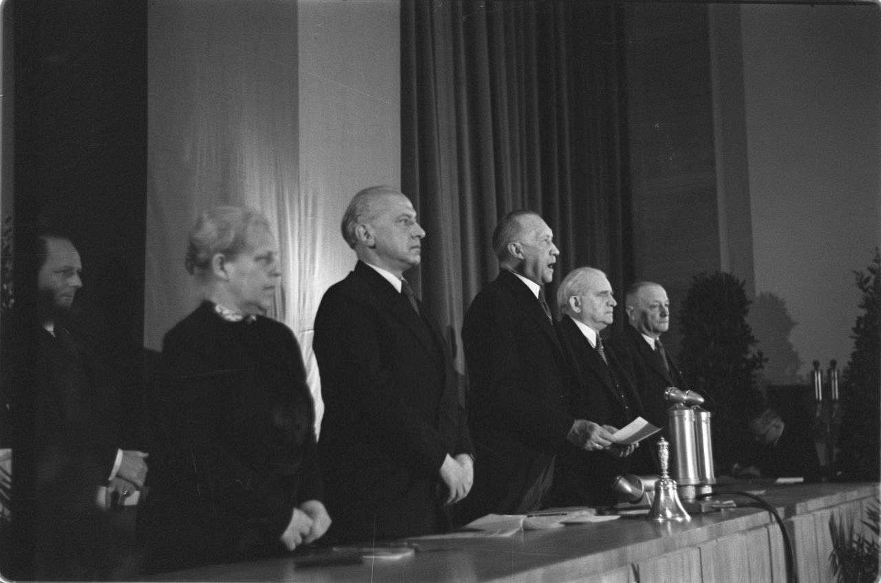 Schwarz-weiße Abbildung von fünf Personen, die nebeneinander hinter einem Tisch stehen. Konrad Adenauer steht sprechend vor Mikrofonen  und hält eine Vorlage in der Hand. Im Hintergrund hängt längs eine deutsche Flagge. Von links nach rechts: Helene Weber, Hermann Schäfer, Konrad Adenauer, Adolf Schönfelder, Jean Stock, Mitglieder des Parlamentarischen Rates.