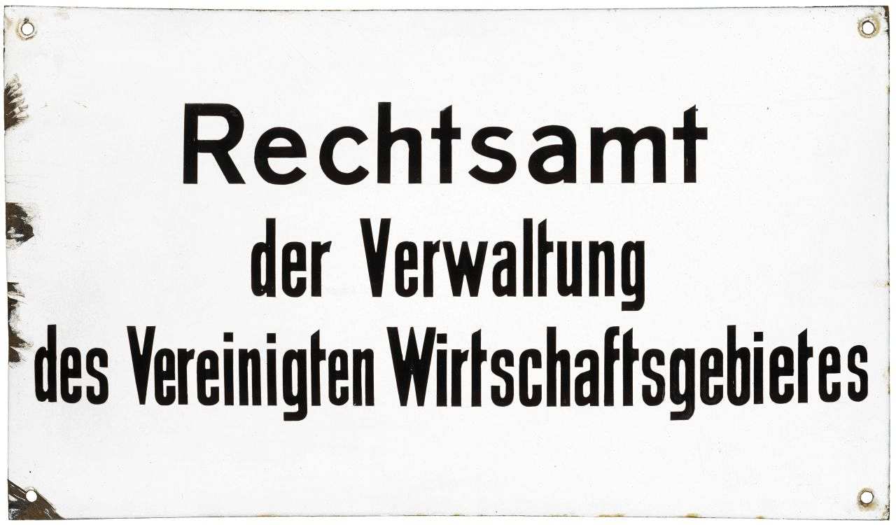 Weißes, gewölbtes Schild; Text (schwarz): Rechtsamt der Verwaltung des Vereinigten Wirtschaftsgebietes. In den Ecken jeweils ein Loch zur Befestigung.