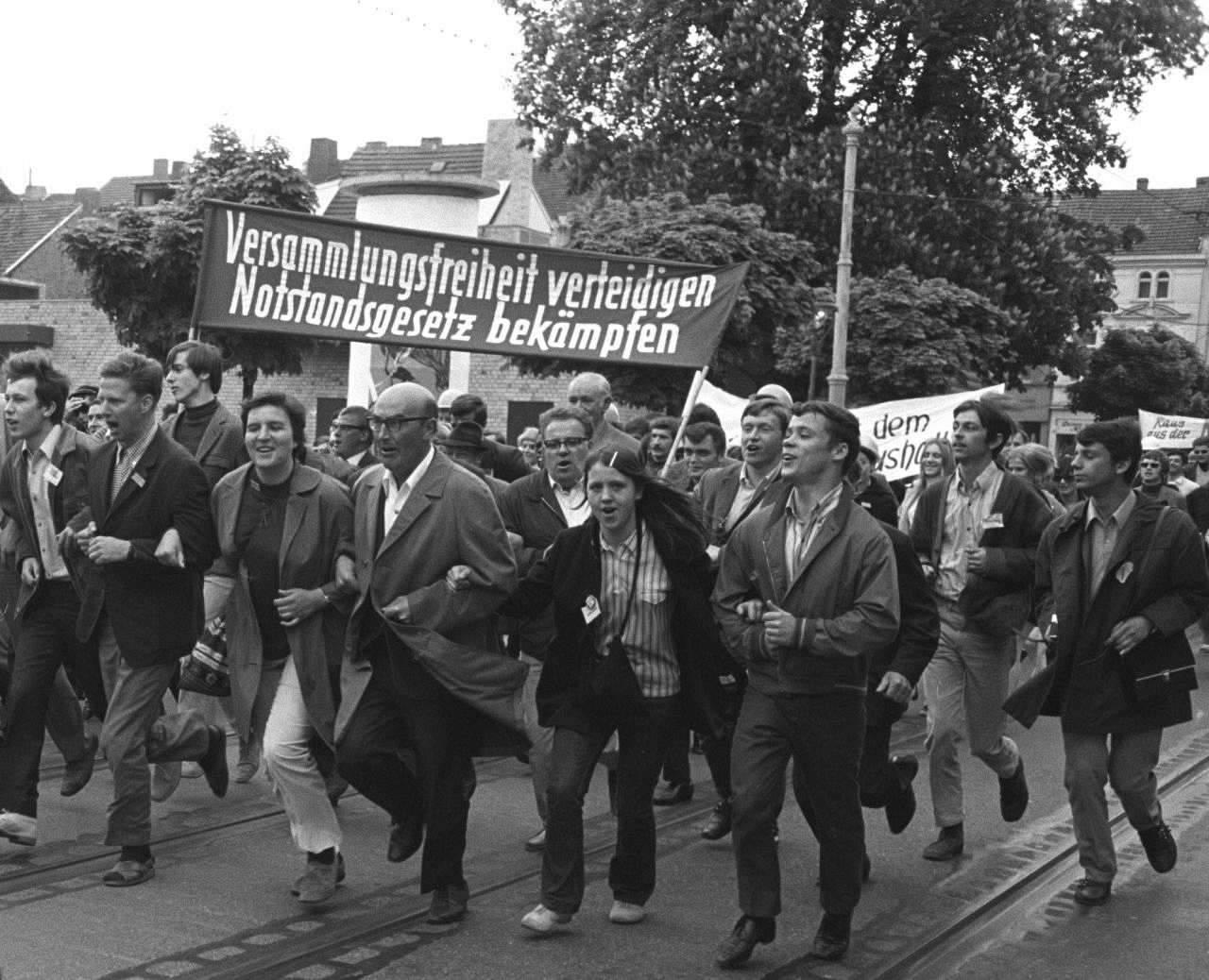 Eine Gruppe von Demonstranten auf einer Straße. Die vier Männer und zwei Frauen in der vordersten Reihe haben sich eingehakt. Hinter ihnen wird ein Spruchband getragen, Aufschrift: Versammlungsfreiheit verteidigen Notstandsgesetz bekämpfen. 