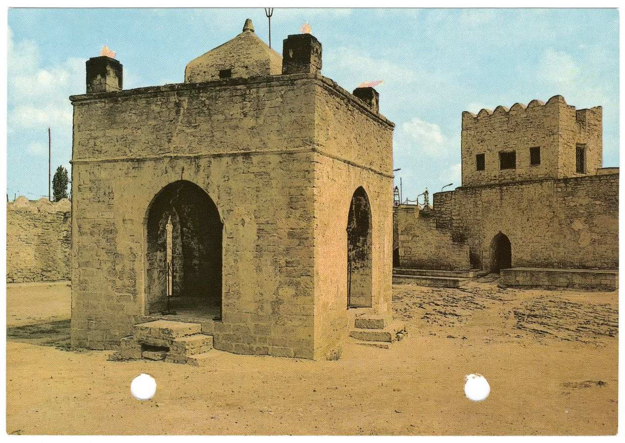 Farbfotografie: Tempel der Feueranbeter im Surachany-Dorf in Baku. Kleines viereckiges Gebäude spitz zulaufenden Eingängen. Galerie mit kleiner Kuppel in der Mitte bilden 1. Stock. Hintergrund: Mauern mit viereckigem Aufbau. Gebäude und Boden sandfarben; blauer Himmel.