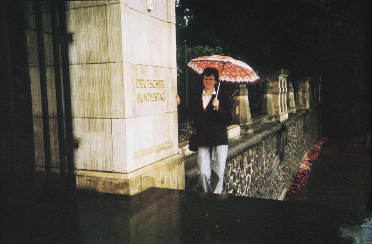 Eine Frau mittleren Alters steht mit Regenschirm auf den Treppenstufen neben dem Eingang des Deutschen Bundestages. Die Bezeichnung steht neben links neben der Frau auf einem Pfeiler.