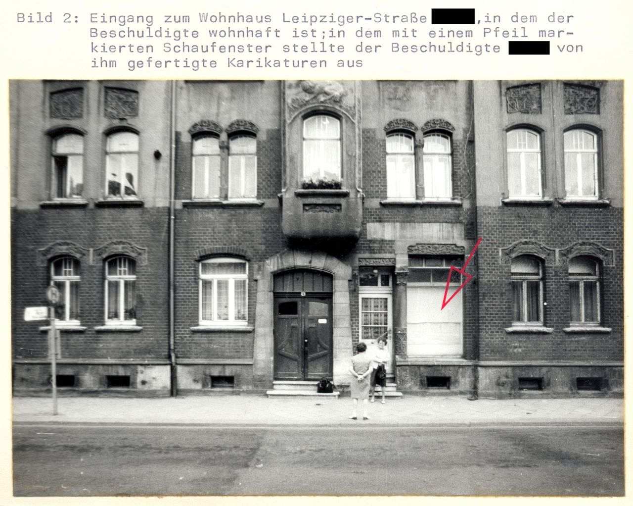 Hausfassade mit Fenstern, Erdgeschoss und erstem Stock. In der Mitte die Eingangstür. Rechts davon ein größeres Fenster, das von innen verhängt ist. Ein in das Bild eingezeichneter roter Pfeil zeigt auf dieses Fenster. Vor der Tür stehen zwei Frauen.