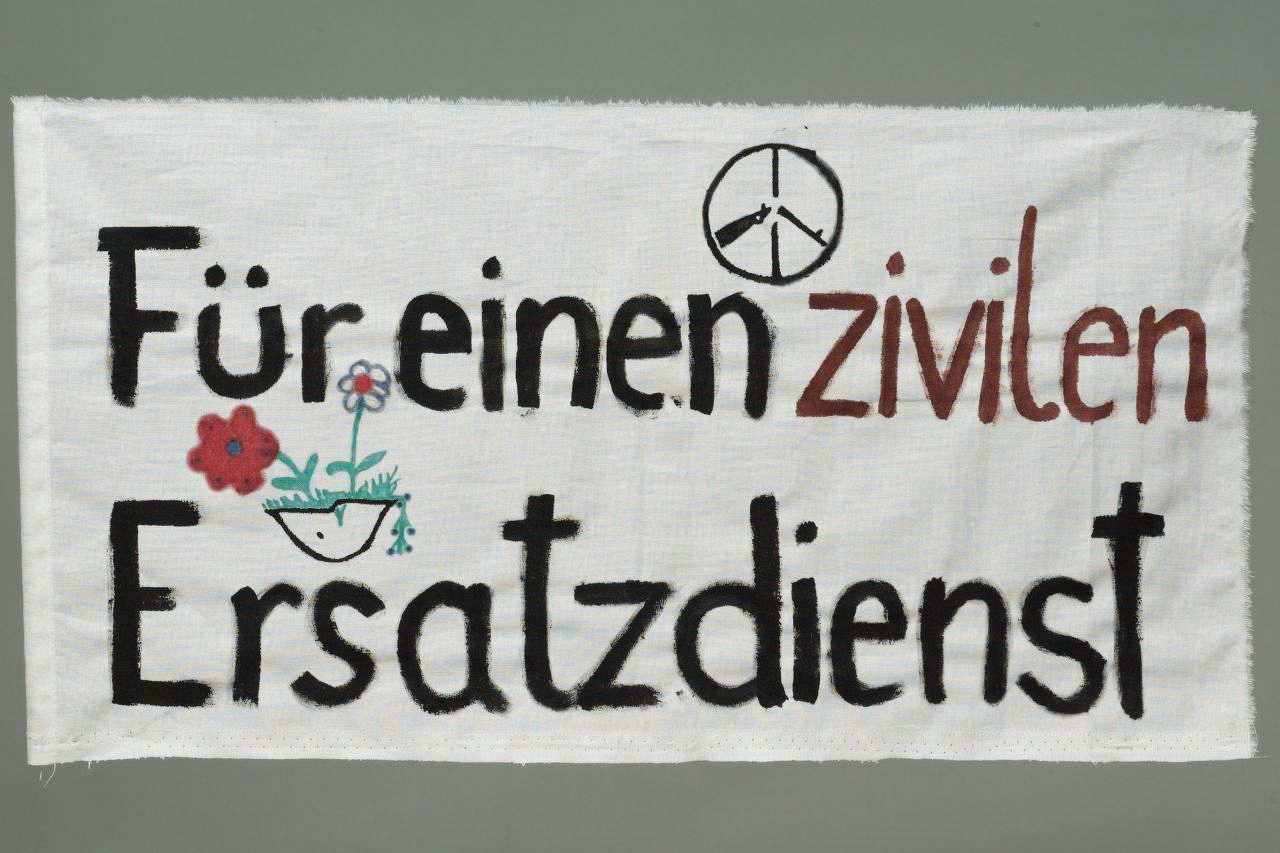 Weiße, querformatige Stoffbahn, schwarz und braun beschriftet. Text: Für einen zivilen Ersatzdienst. Oben das Symbol der internationalen Friedensbewegung (Kreis mit durchgebrochenem Gewehr) und links mittig Blumen, die aus einem Stahlhelm sprießen.