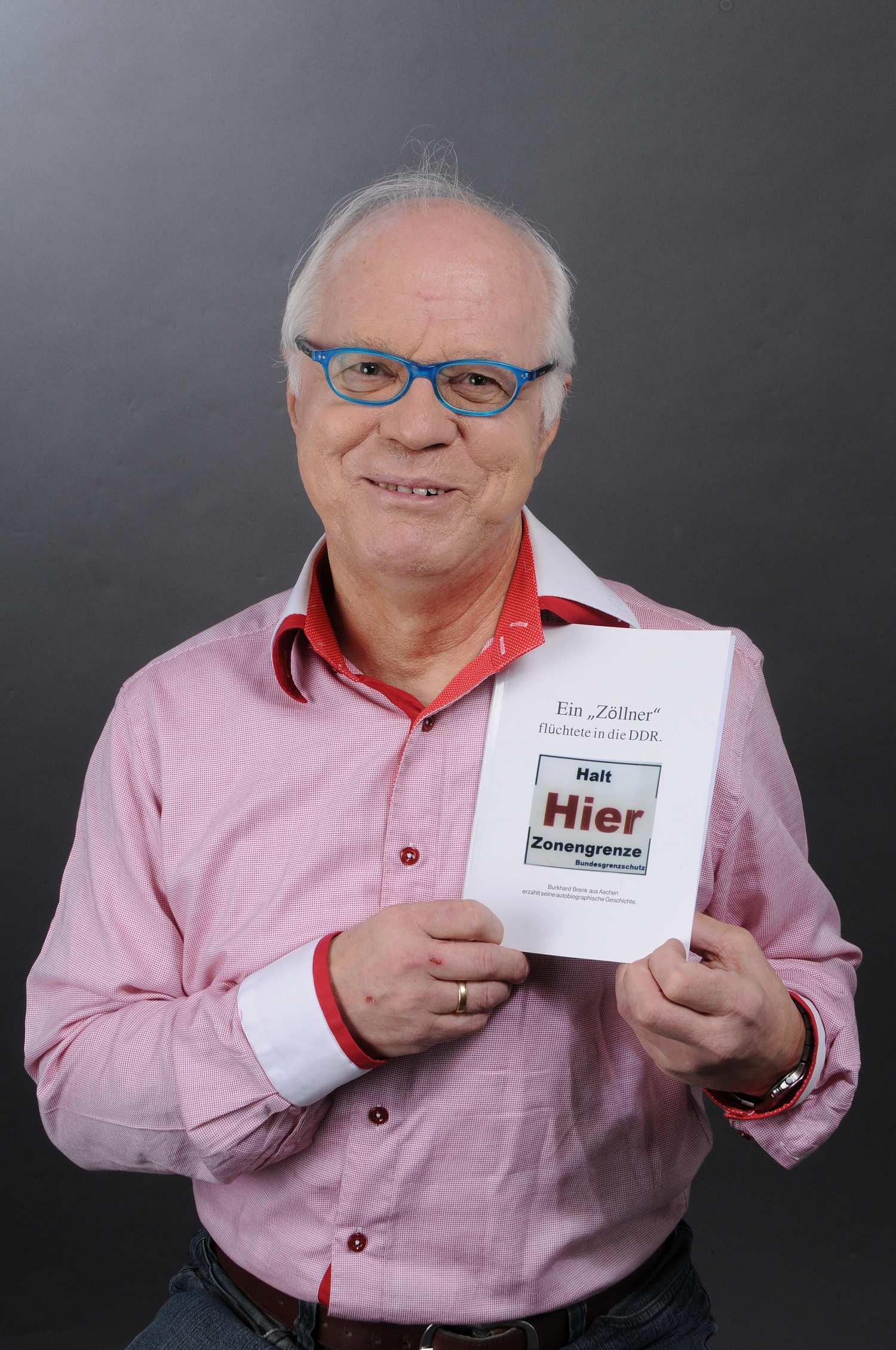 Porträtfoto von Zeitzeuge Burkhard Brenk mit blauer Brille und Buch in der Hand, 2016.