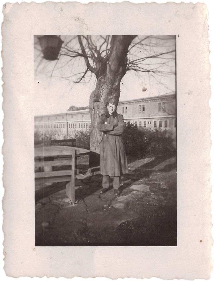 Fotografie, schwarzweiß. Ein junger Mann steht unter einem Baum. Er trägt eine Wehrmachts-Uniform mit Stiefeln, eine schräg aufgesetzte Feldmütze und eine Brille. Links neben ihm befindet sich eine Sitzgruppe aus Holz. Im Hintergrund erkennt man Kasernengebäude.