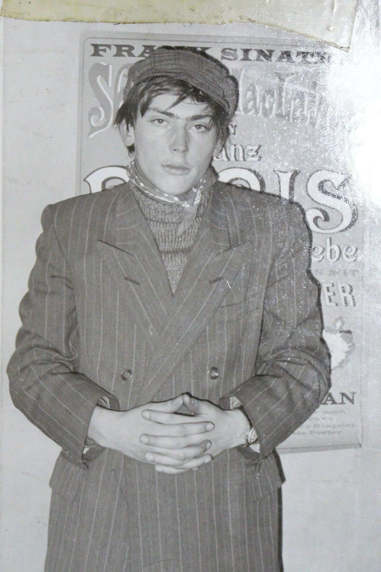 Ein junger Mann mit längeren Haaren steht mit gekreutzen Händen vor einem Musikplakat. Er trägt ein Nadelstreifen-Jacket und eine Kappe.