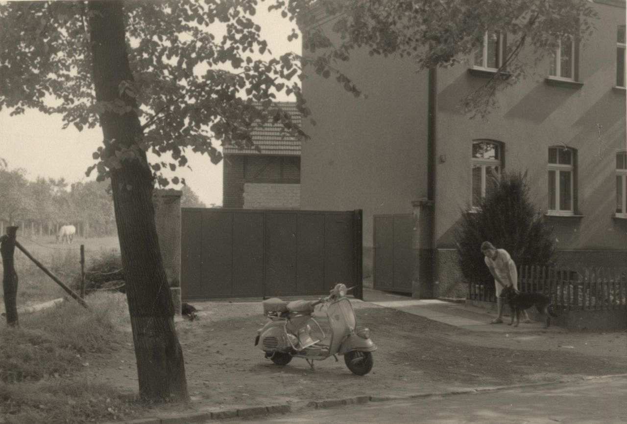 Schwarz-weiß-Fotografie: Eine Vespa steht mittig im Bild, neben einem Baum, vor einem offenen Tor, das zu einem Wohnhaus im Hintergrund führt. Außerdem rechts im Bild eine Frau mit einem Hund.