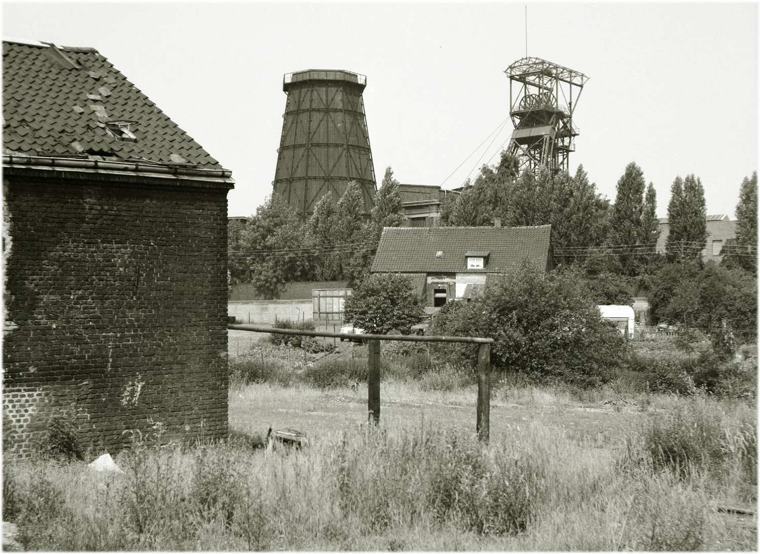 Fotografie, schwarzweiß. Landschaftsaufnahme eines verwilderten Gartens, am linken Bildrand steht ein großes Backsteinhaus. In der Mitte des Bildes befindet sich ein Einfamilienhaus. Dahinter ragen ein Förderturm und andere Industrieanlagen empor.