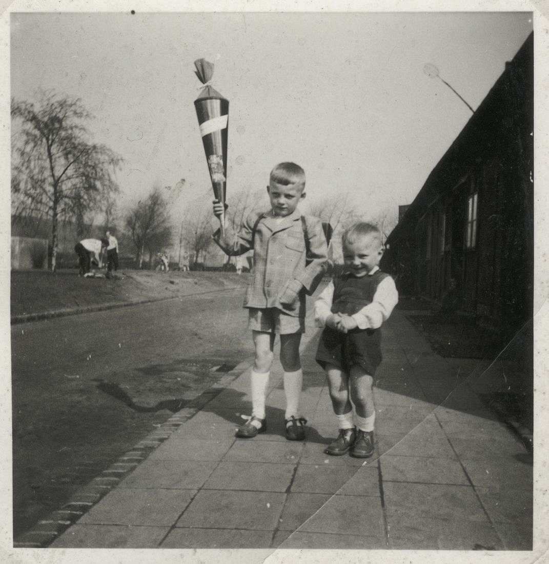 Fotografie, schwarzweiß. Zwei Jungen stehen auf einem Gehweg. Beide schauen in die Kamera Der linke, ältere Junge hält eine Schultüte hoch und schaut ernst, während der jüngere, rechte Junge kichert. 
