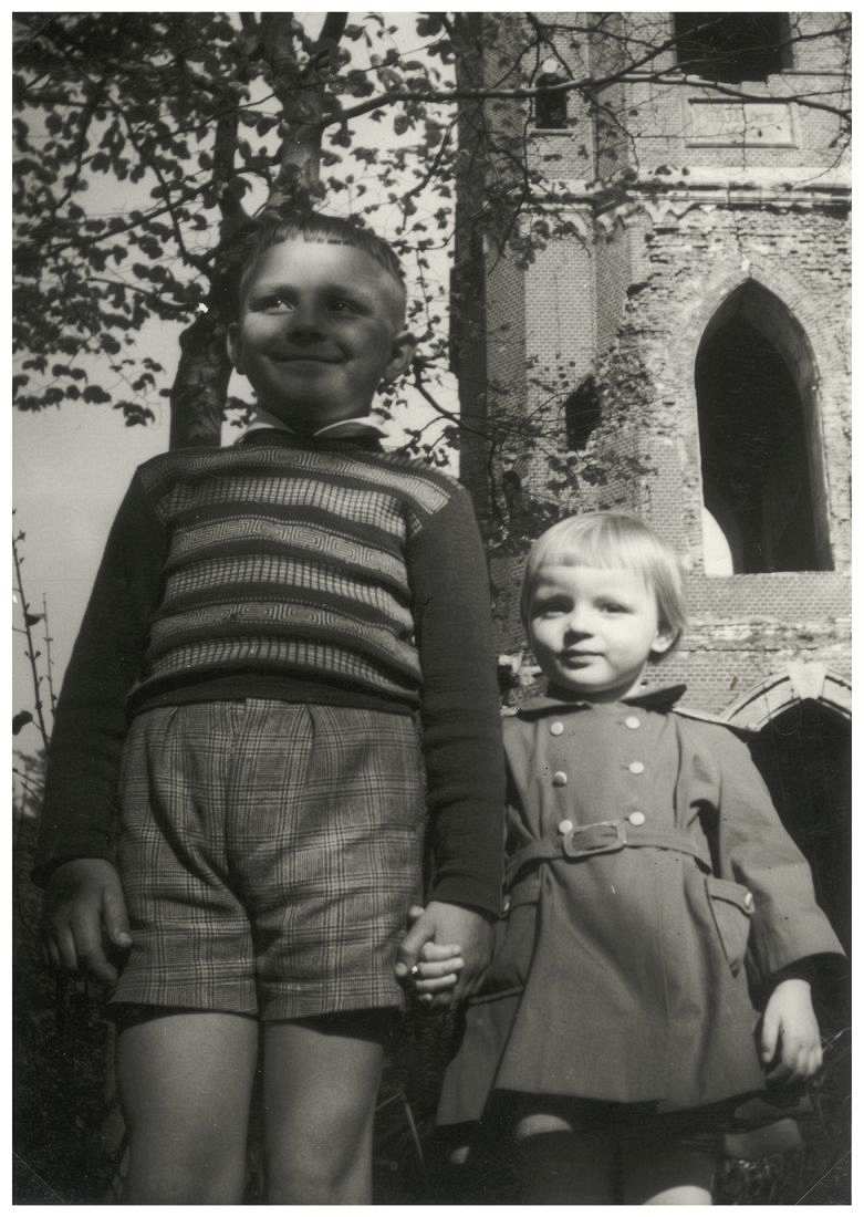 Fotografie, schwarzweiß. Ein Junge steht im Vordergrund und blickt stolz nach oben. Er hat ein jüngeres Mädchen an der Hand, das in die Kamera blickt. Im Hintergrund ist ein Turmruine zu sehen.