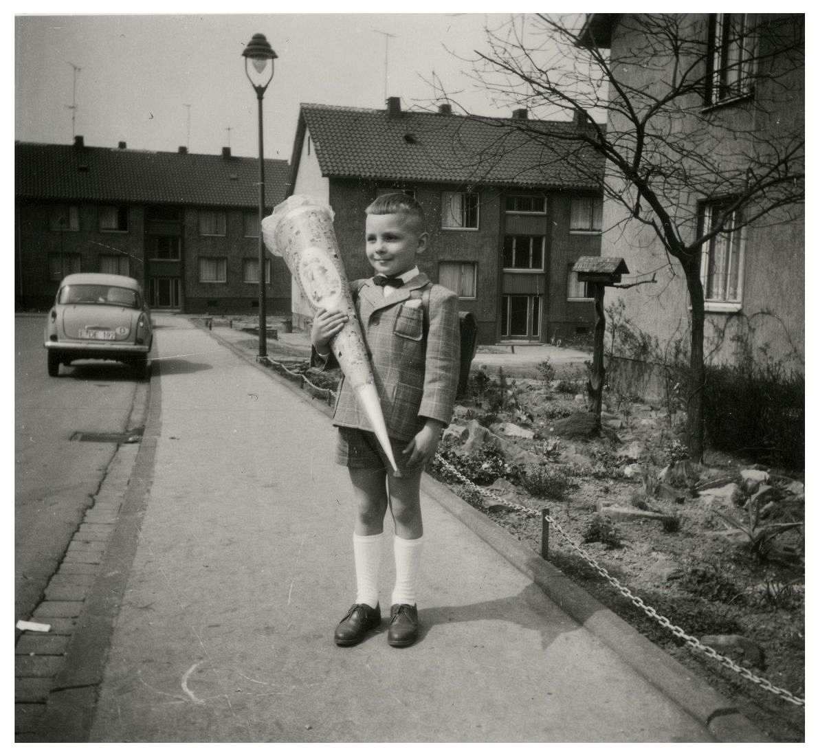 Fotografie, schwarzweiß. Ein Junge steht auf einem Gehweg. Er trägt einen Schulranzen und hält eine Schultüte in der Hand. Links neben ihm parkt ein Kleinwagen am Straßenrand.