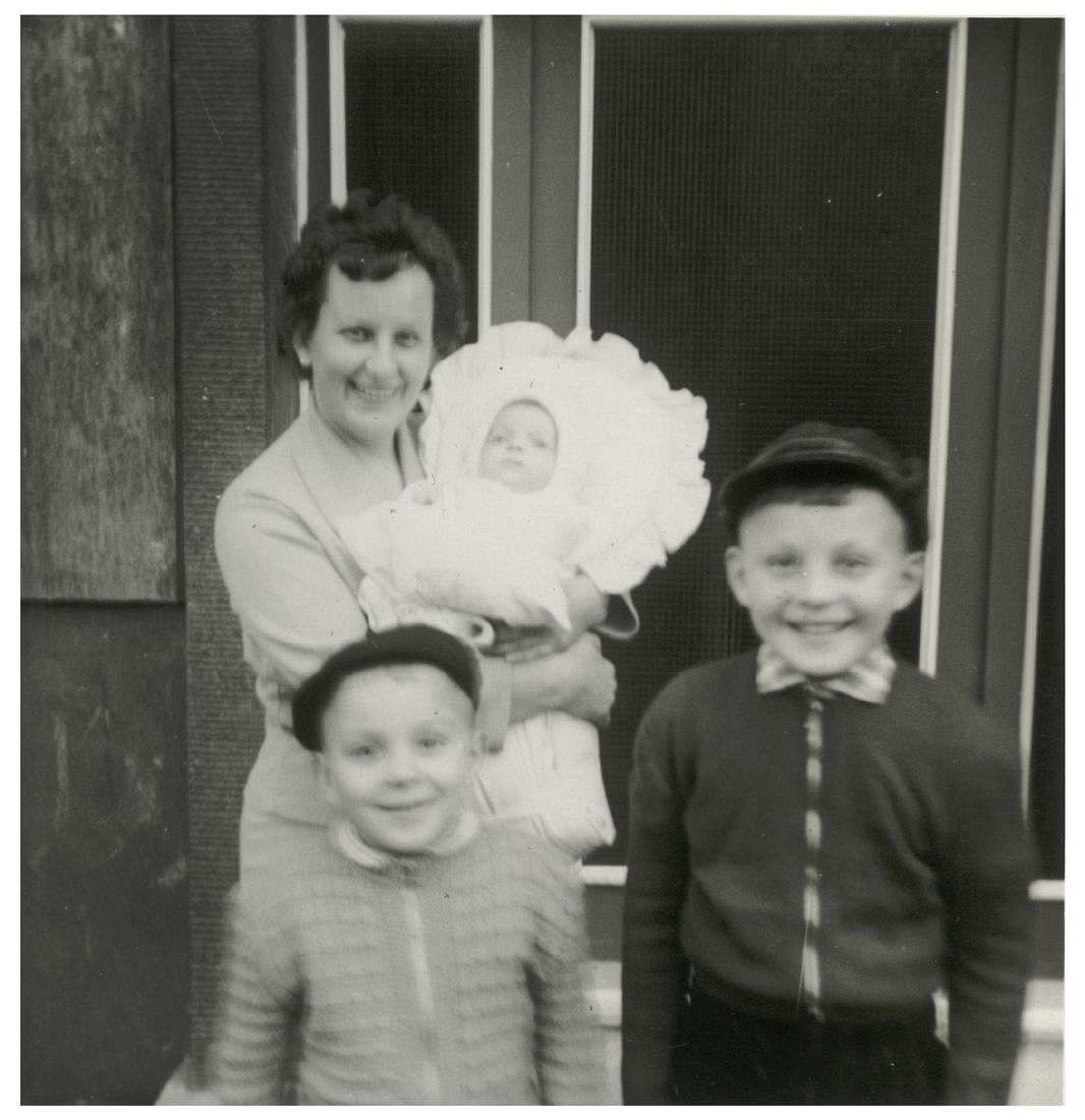 Fotografie, schwarzweiß. Eine Frau steht vor einem Hauseingang. Auf dem Arm hat sie ein Kleinkind. Vor ihr stehen zwei Jungen, die in die Kamera lächeln. 