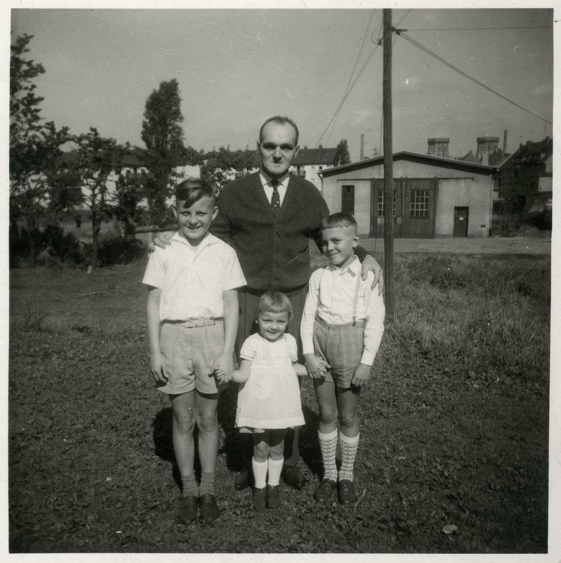 Fotografie, schwarzweiß. Zwei Jungen stehen auf einer Wiese. Zwischen ihnen steht ein jüngeres Mädchen, das von den beiden Jungen an den Händen gehalten wird. Hinter den Kindern steht ein Mann, der mit seinen Armen die Kinder einrahmt. 