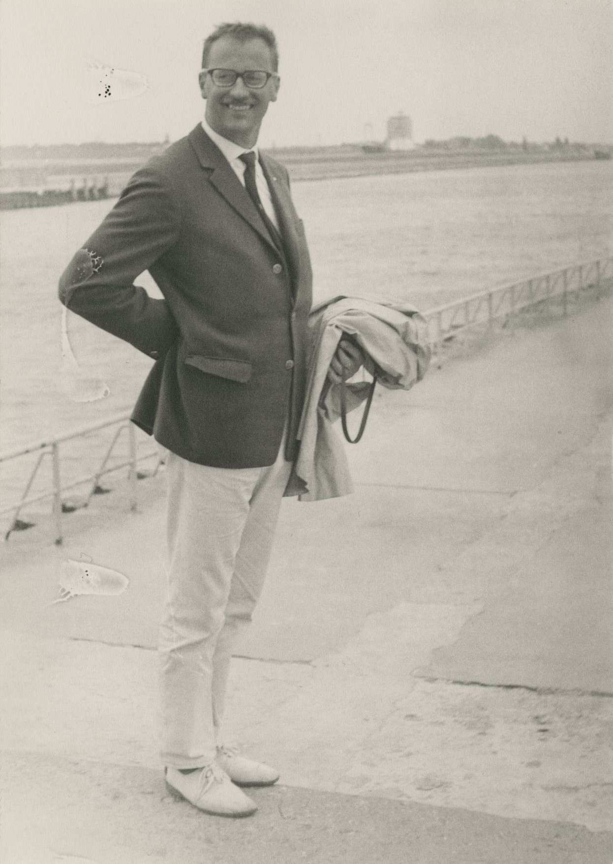 Foto von Lutz Rackow, ca. 38 Jahre alt. Er steht auf einer Hafenpromenade und trägt eine sportliche Kombination aus dunklem Sakko, heller Hose. Über einem angewinkelten Arm hängt eine helle Jacke. Im Hintergrund erkennt man ein Gewässer und Schiffsanleger.