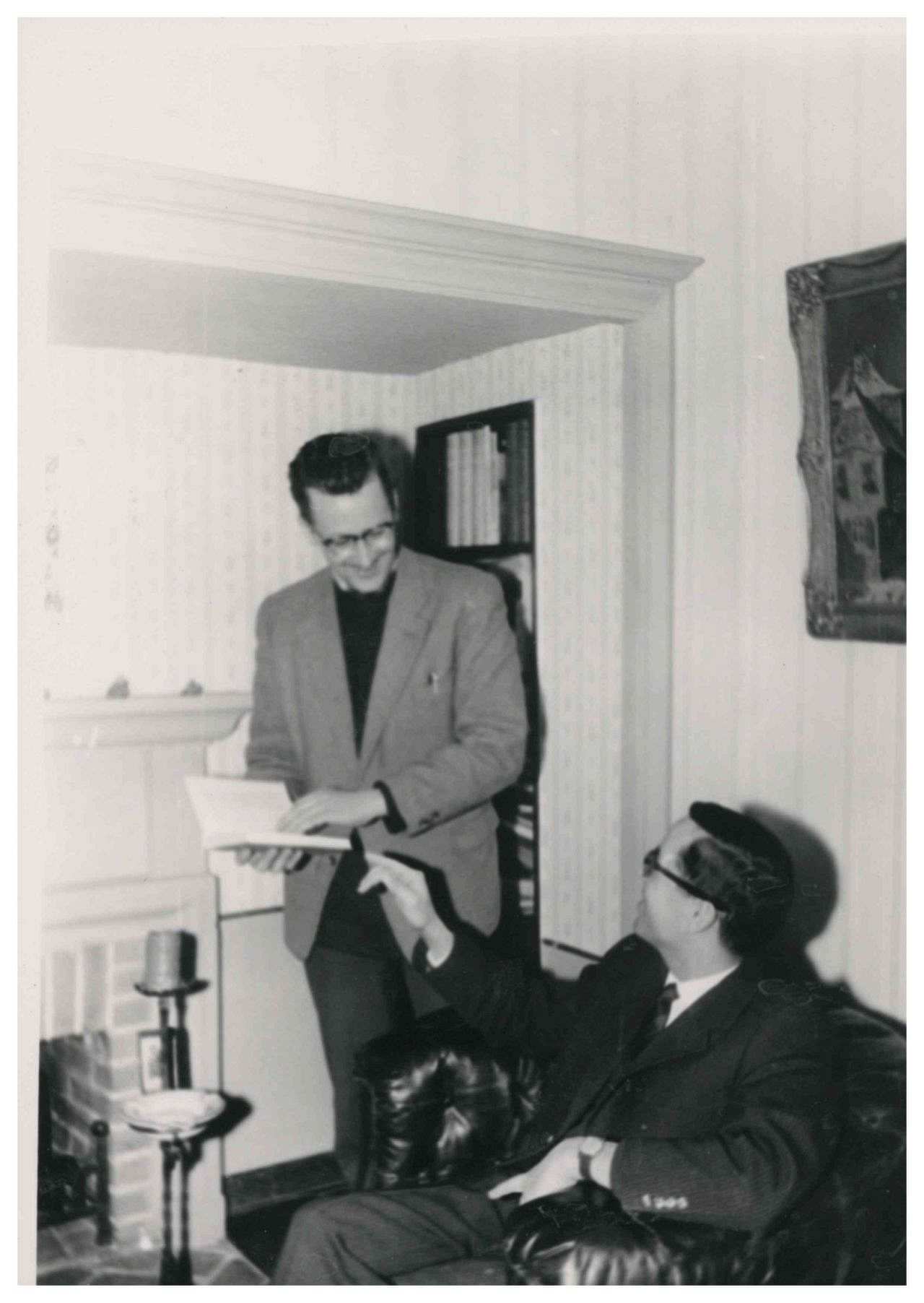 Foto, schwarz-weiß. Zwei Männer in einem Wohnzimmer. Der eine Mann sitzt in einem Er schaut den anderen Mann an, der links neben ihm steht und ein Buch aufgeschlagen hat. Im Hintergrund erkennt man ein Bücherregal und einen Kamin.