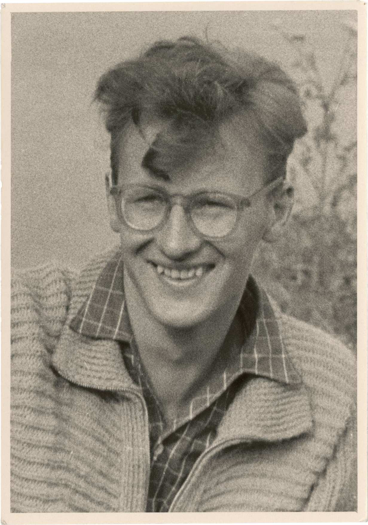 Porträtfoto von Lutz Rackow, ca. 23 jahre alt. Er trägt ein karriertes Hemd, mit einer Wolljacke darüber und eine Brille. Die lockigen, mittellangen Haare fallen teilweise ins Gesicht. Er schaut leicht an der Kamera vorbei und lächelt.