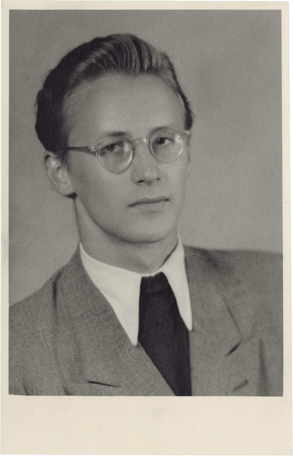 Porträt-Foto von Lutz Rackow, ca. 19 Jahre alt, vor grauem Hintergrund. Er trägt einen Anzug mit Pulli und eine Brille. Die Haare mittellangen Haare sind zurückgekämmt. Er ist leicht seitlich aufgenommen und schaut direkt in die Kamera. 