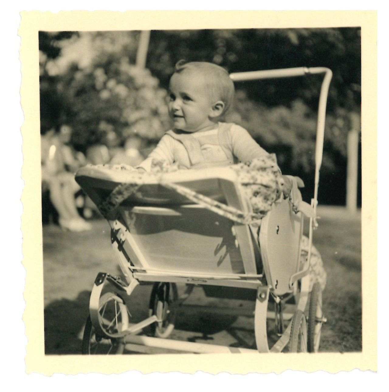 Quadratische schwarz-weiß Fotografie, ein einjähriges Baby sitzt aufrecht in einem offenen Kinderwagen und schaut aufmerksam hoch.