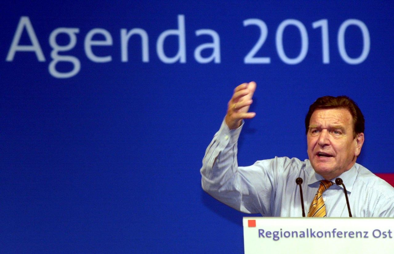 Bundeskanzler Gerhard Schröder hält engagiert eine Rede. Er ist rechts unten im Bild zu sehen, trägt Hemd und Krawatte. Seine rechte Hand ist in einer Redegeste erhoben. Dominiert wird das Foto von der blauen Wand im Hintergrund, auf der groß in hellblau die Beschriftung Agenda 2010 zu lesen ist.