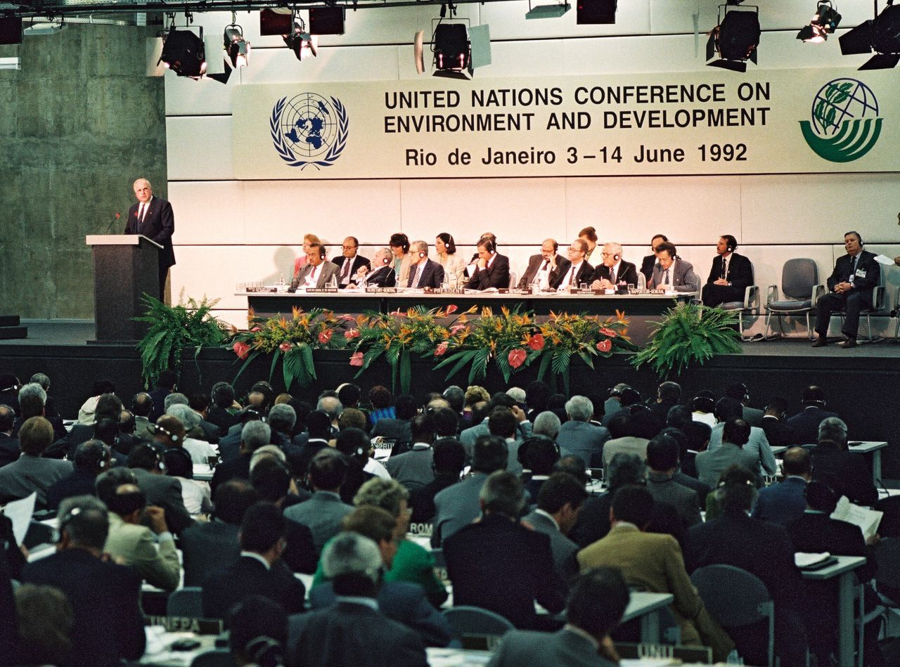 Farb-Foto, Helmut Kohl hinter Redner-Pult auf einer Tribüne, rechts davon Tisch mit weiteren Delegierten. Hinter ihnen ein Schild, darauf: ‚United Nations Conference on Environment and Development, Rio de Janeiro 3-14 June 1992‘. Publikum sitzt an Tischen.