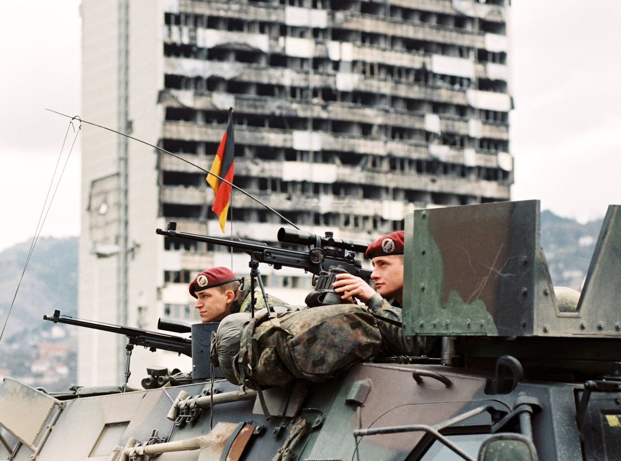Farbfoto, ein Ausguck eines Militärfahrzeugs, aus dem zwei Soldaten Ausschau halten, mit jeweils einem Gewehr in Bereitschaft. Ein Soldat hält ein Fernglas bereit. Hinter ihnen an einem Stab die deutsche Flagge. Im Hintergrund schwer beschädigtes Hochhaus.