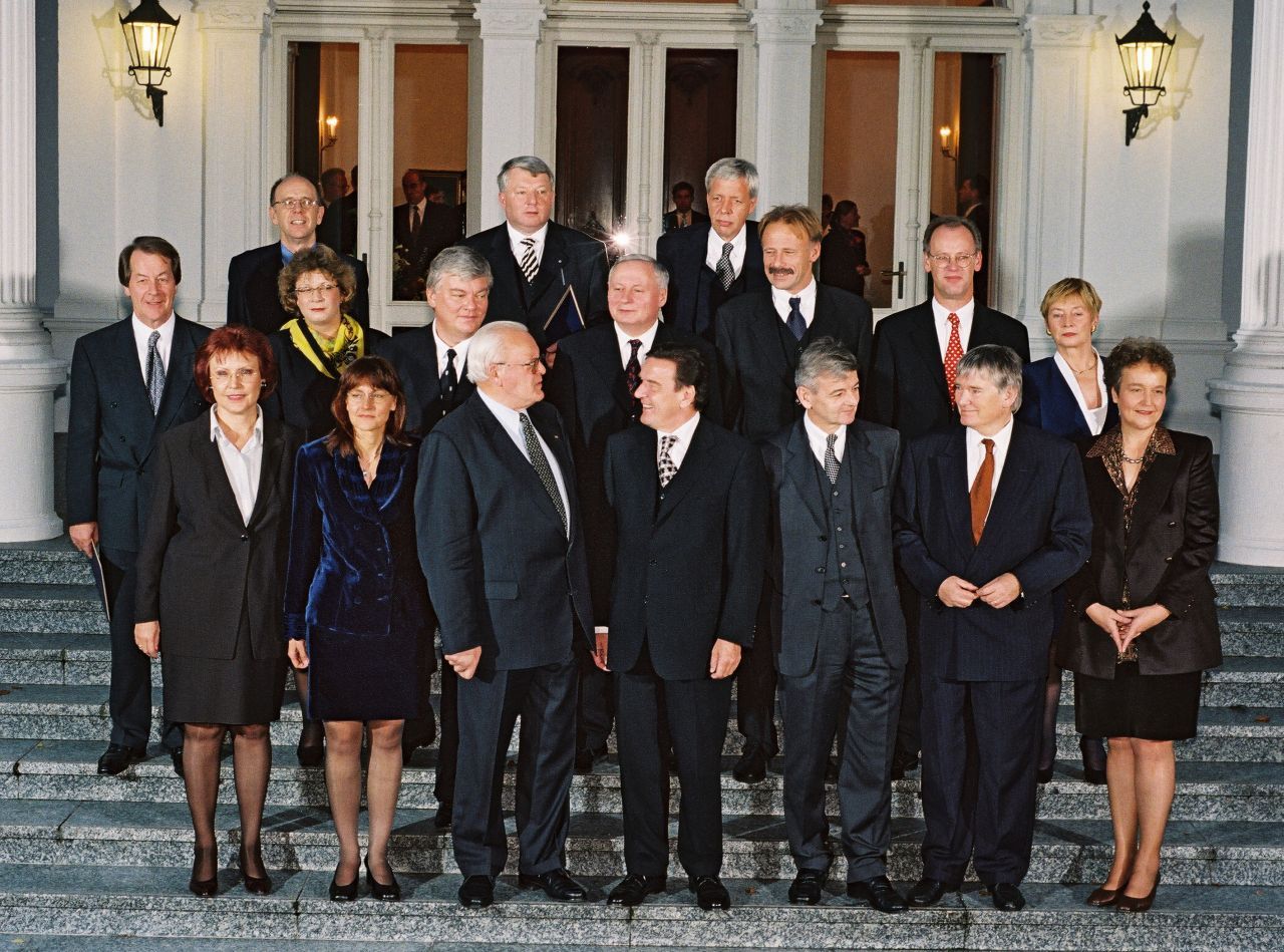 Farbfoto, auf einer Treppe vor der Villa Hammerschmidt in drei Reihen aufgestellt die Mitglieder des Kabinetts Schröder mit dem Bundespräsidenten Roman Herzog. Im Hintergrund Fensterfront des Gebäudes, flankiert von zwei leuchtenden Wandlaternen.