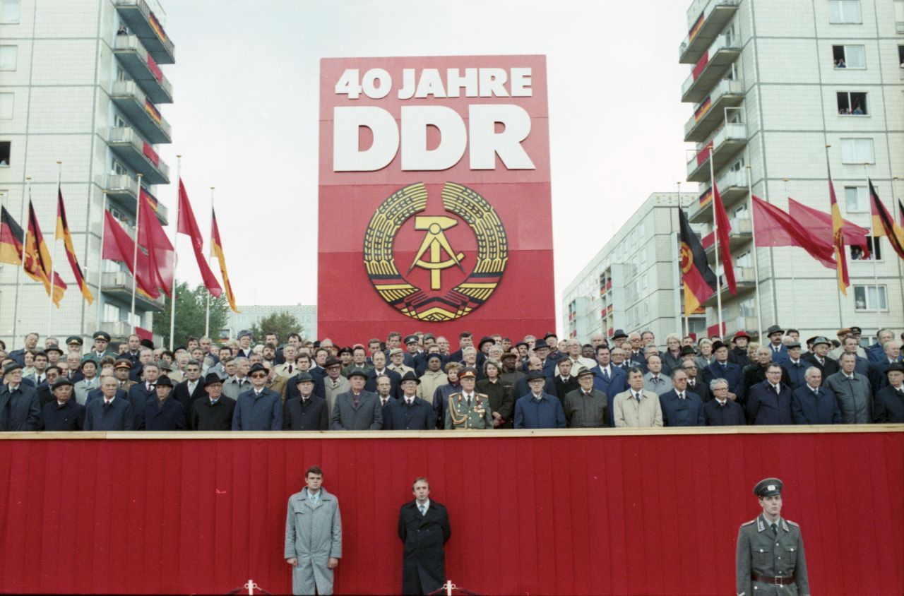 Auf dem Bild ist die Ehrentribüne zu sehen während der Ehrenparade der Nationalen Volksarmee auf der Feier des 40. Jahrestags der DDR. Die wichtigen Gäste stehen auf einer rotumkleideten Tribüne zwischen zwei Hochhäusern. Im Hintergrund ist ein sehr großes, ca. 10m hohes Plakat zu sehen mit der Aufschrift: 40 Jahre DDR und dem Staatswappen. Außerdem: Mehrere rote Fahnen und DDR-Flaggen.