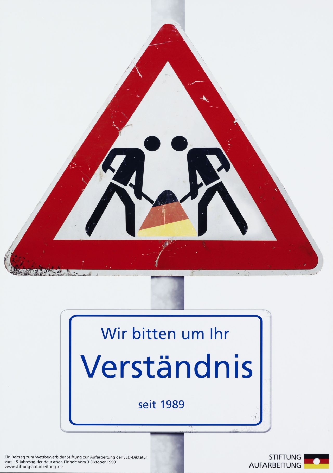 Farbiges Plakat; weißer Hintergrund; dreieckiges Baustellenschild: zwei Strichmännchen mir Schaufeln arbeiten gemeinsam an einem schwarz-rot-goldenen Sandhaufen; unterhalb des Schildes ist weißes Schild mit blauer Schrift angebracht: Wir bitten um Ihr / Verständnis / seit 1989; unten, links, in schwarzer Schrift:  Ein Beitrag zum Wettbewerb der Stiftung zur Aufarbeitung der SED-Diktatur / zum 15. Jahrestag der deutschen Einheit vom 3. Oktober 1990 / www.stiftung-aufarbeitung.de; daneben, rechts: Stiftung / Aufarbeitung, mit Logo.